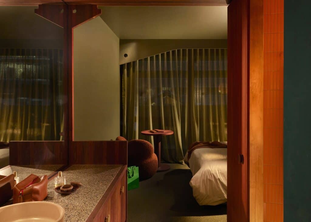 Olive-toned guest room at Melbourne Place Hotel seen past a terrazzo vanity and jarrah mirror frame, with green curtains and a round side table
