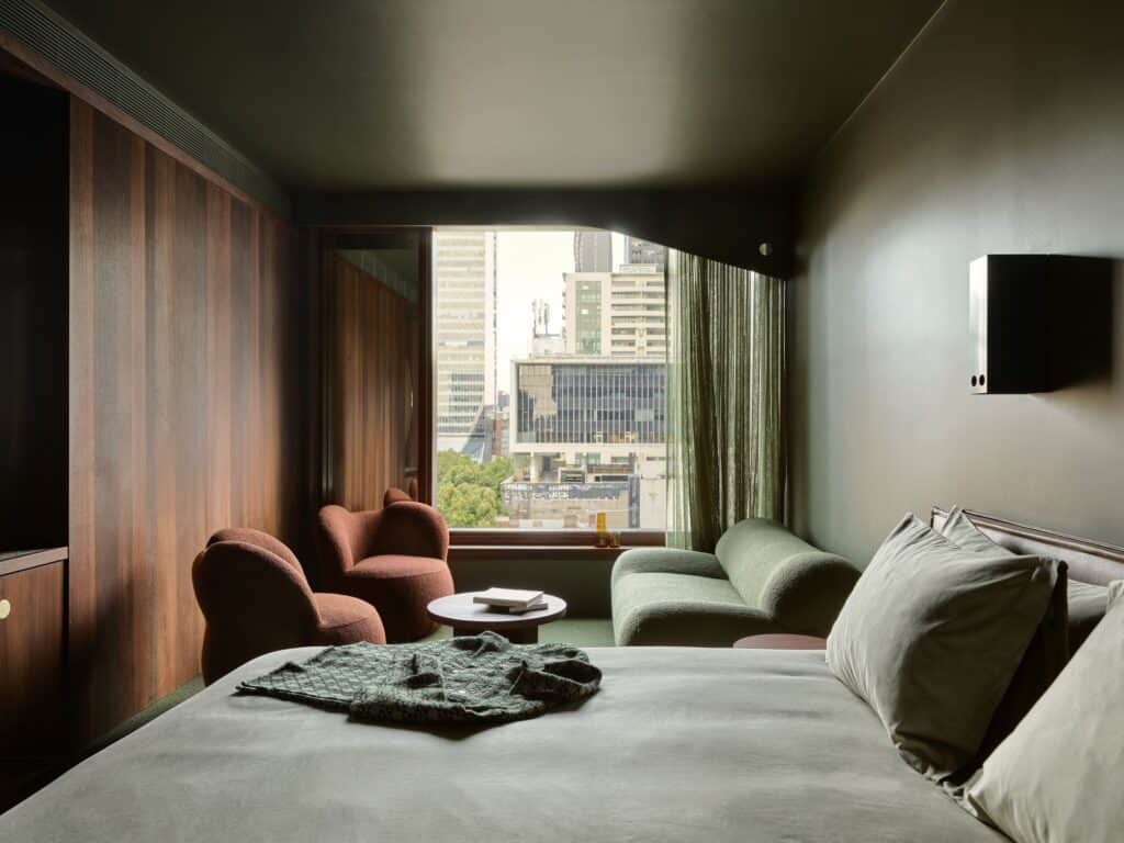 Deep green guest room at Melbourne Place Hotel with jarrah wall panelling, two rust boucle chairs, a green sofa and a window onto city towers
