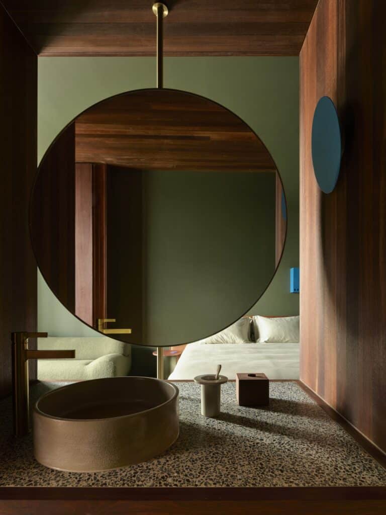 Round mirror hung from a brass rod above a ceramic basin and terrazzo counter in a green guest room at Melbourne Place Hotel