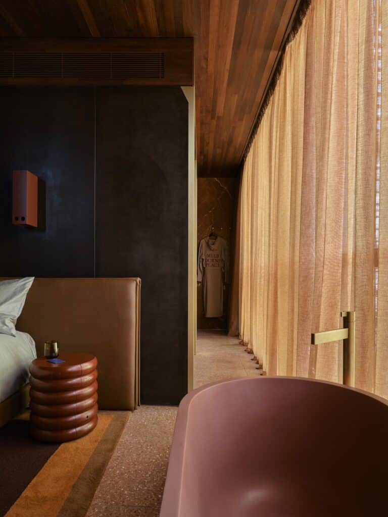 Dusty pink bathtub beside full-height linen curtains in a dark corten-walled suite at Melbourne Place Hotel, with a leather headboard