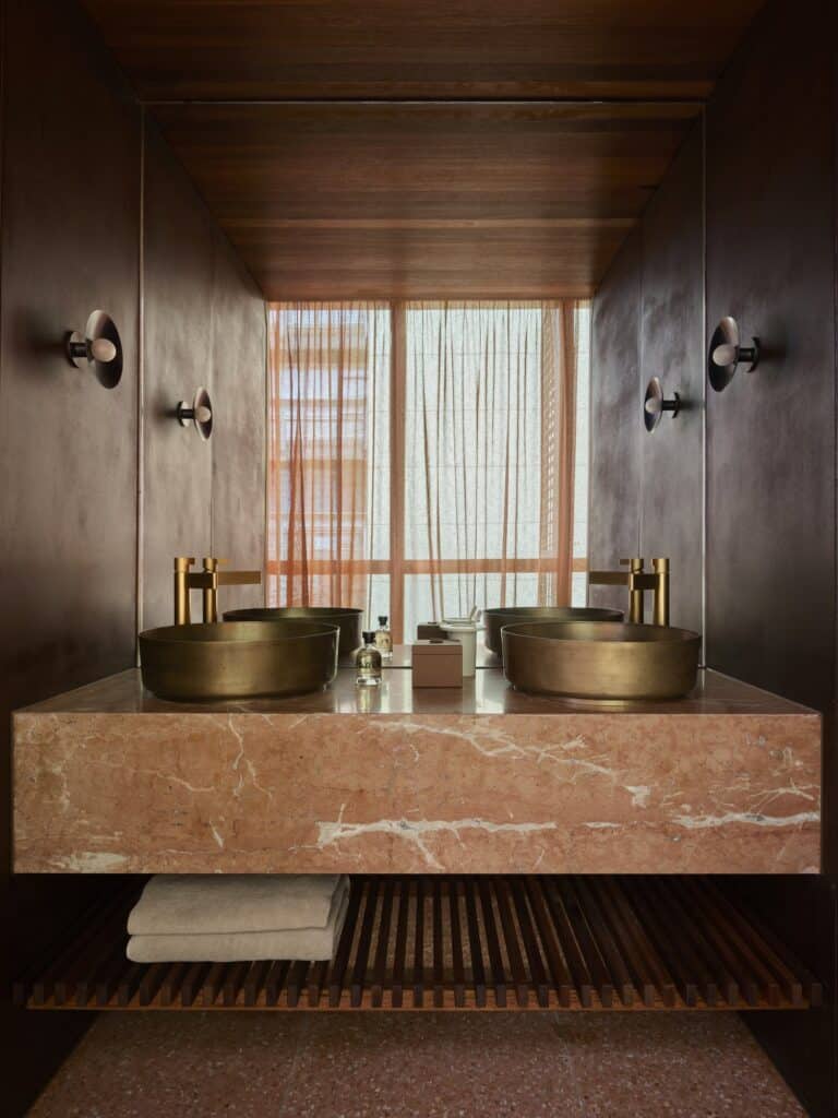 Rose marble double vanity with two brass vessel basins and a slatted jarrah shelf below, framed by mirrors at Melbourne Place Hotel