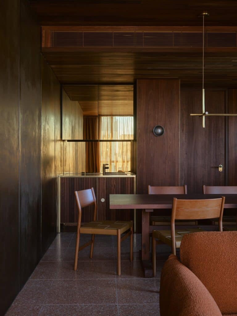 Jarrah-panelled suite at Melbourne Place Hotel with terrazzo floor, timber dining chairs with leather seats, a brass pendant and kitchenette