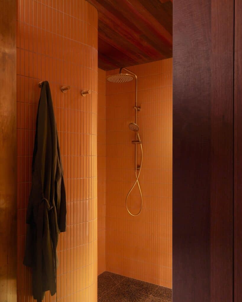 Shower at Melbourne Place Hotel lined in orange vertical tiles with brass rain shower fittings, a dark robe hanging on a hook beside it