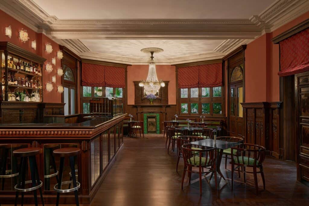 The Caffè at Mi Shang at Prada Rong Zhai in Shanghai, a long carved wooden bar with a glass-fronted counter and leather stools, terracotta walls with glowing sconces, red Roman blinds, a crystal chandelier and bentwood chairs around marble tables