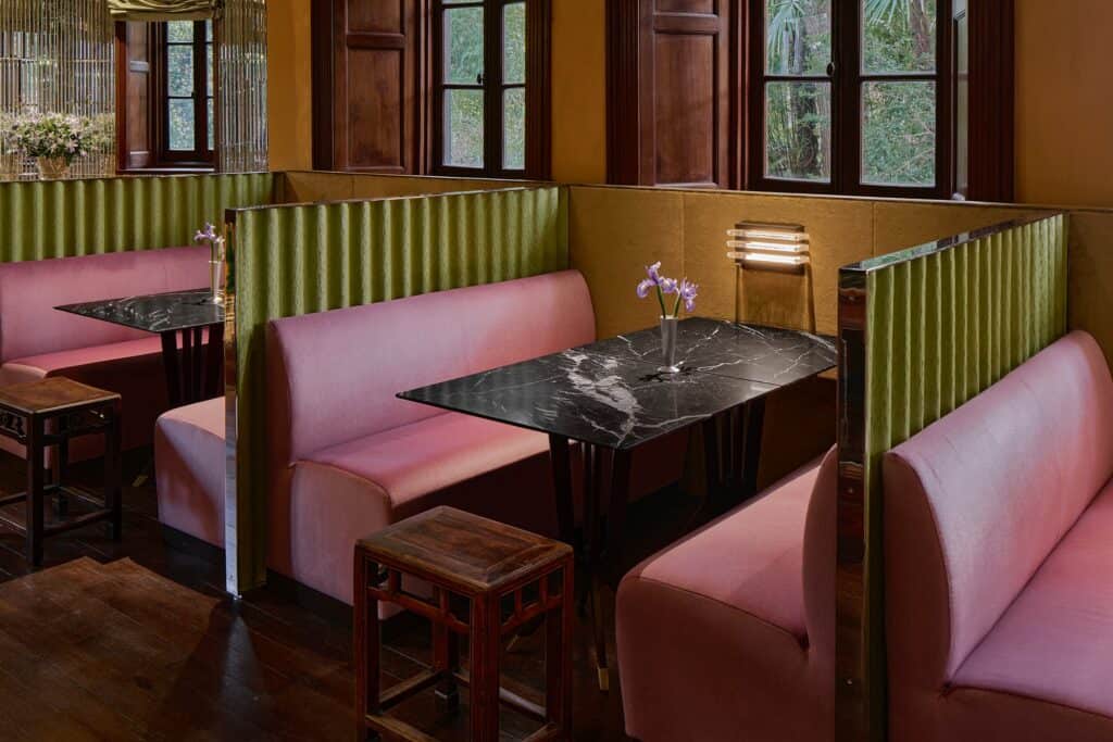 Dining booths at Mi Shang at Prada Rong Zhai, dusty pink velvet banquettes divided by pleated olive-green screens with mirrored edges, black marble tables with single irises and carved Chinese wooden stools on a dark timber floor