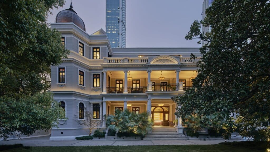 Rong Zhai at dusk, the restored 1918 villa housing Mi Shang at Prada Rong Zhai, a grey stucco facade with a copper-domed corner tower, columned loggias glowing with warm light, palms and magnolias in the garden and a glass tower behind