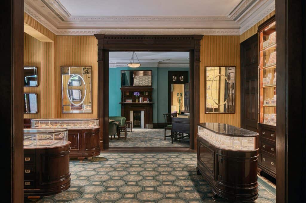 Pastry Shop at Mi Shang at Prada Rong Zhai, curved dark wood and glass display counters on a patterned blue-green floor, striped wallpaper with mirrors and a lit cabinet of boxed pastries, a doorway framing a teal room beyond