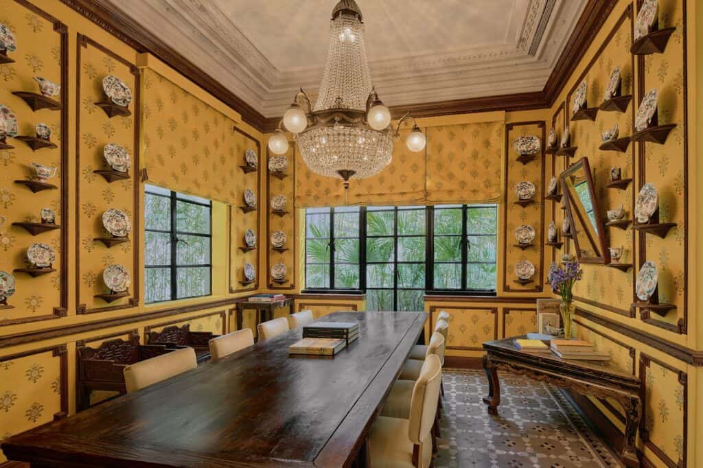 The Study at Mi Shang at Prada Rong Zhai, a long dark wooden table with cream chairs under a crystal chandelier, mustard patterned walls lined with porcelain plates on small brackets and a black steel window onto palms