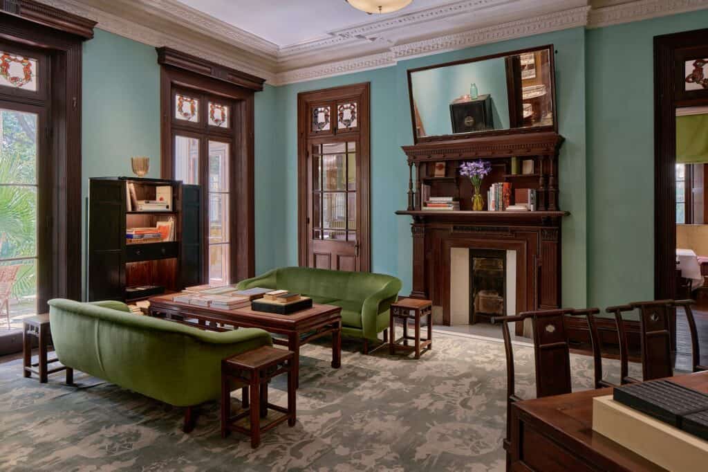 The Library at Mi Shang at Prada Rong Zhai, two curved olive-green velvet sofas around a low Chinese wooden table stacked with books, teal walls, a carved overmantel mirror and tall doors with painted glass panels