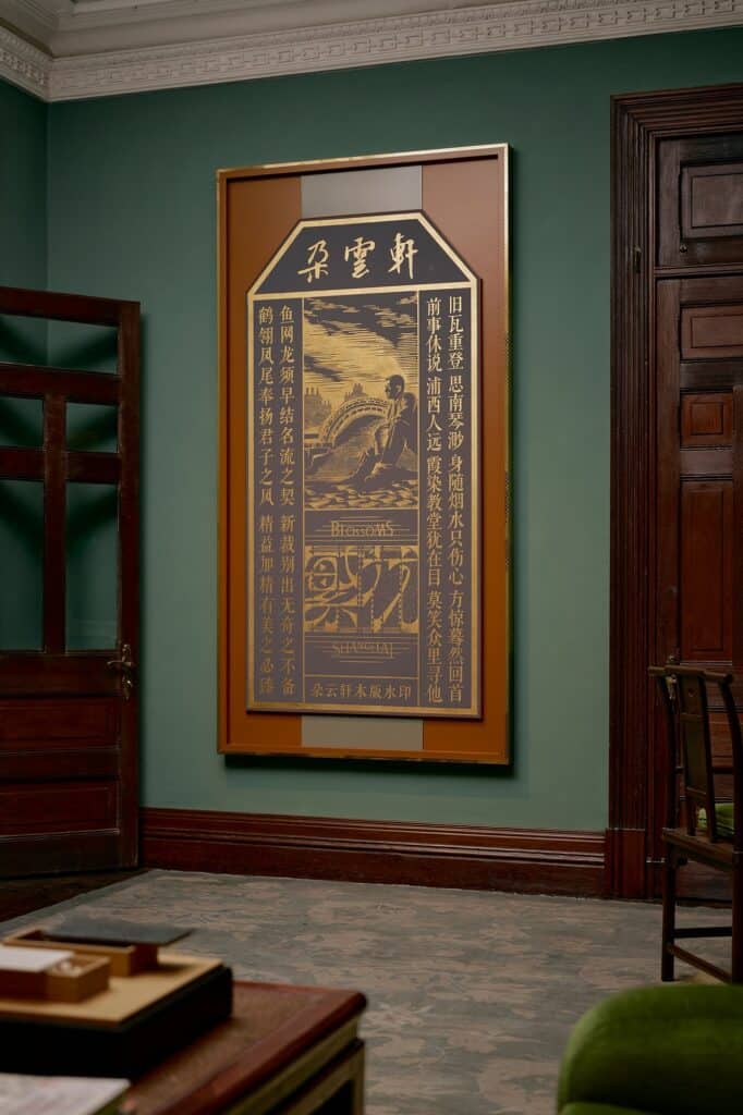 Framed gold and black woodblock print from Wong Kar Wai's Rising Clouds, Blooming Flowers series with Duo Yun Xuan at Mi Shang at Prada Rong Zhai, Chinese calligraphy around a seated figure and the words Blossoms Shanghai, on a green wall beside a dark wooden door
