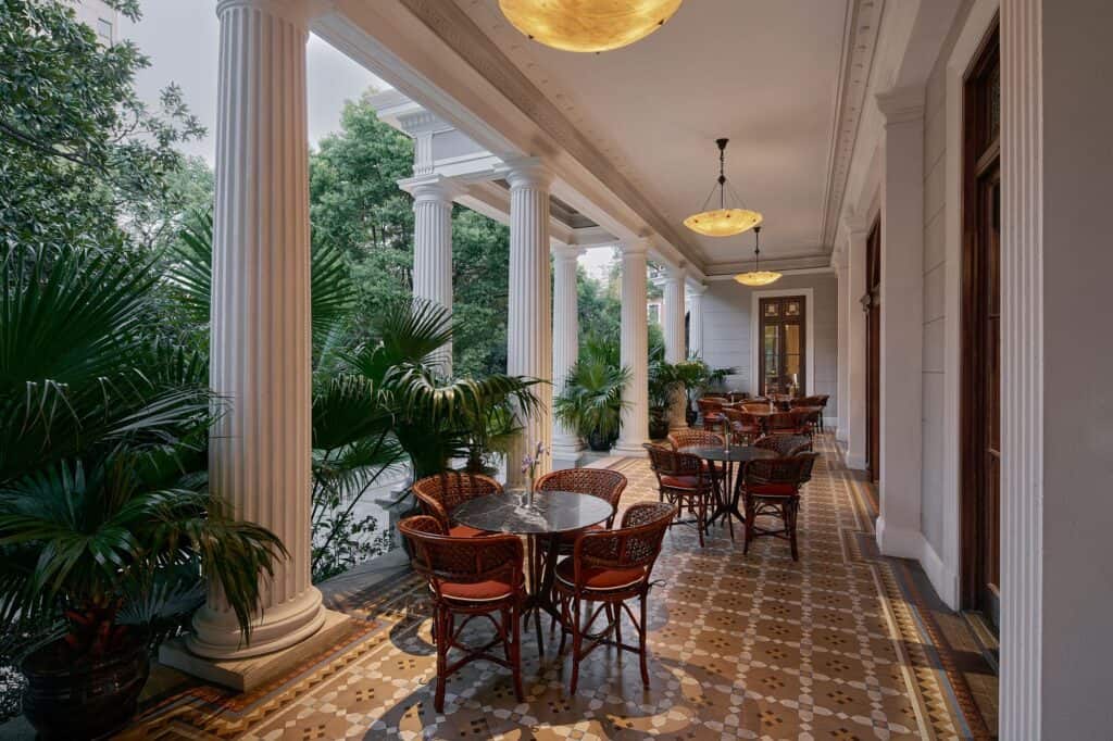 The Terrace at Mi Shang at Prada Rong Zhai, a colonnade of white fluted columns with bamboo and rattan chairs around round stone tables on a patterned tile floor, alabaster pendant lights and potted palms