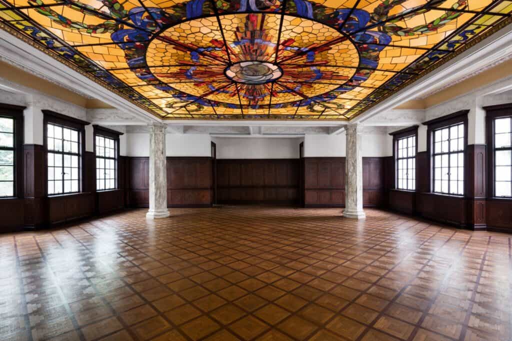 Ballroom of Prada Rong Zhai in Shanghai, a vast stained-glass skylight in amber and blue above a pale room with dark wood wainscoting, marble columns and a polished parquet floor laid in a diagonal pattern