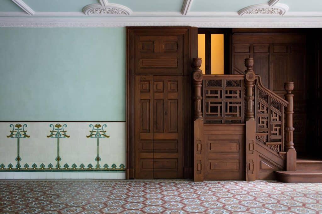 Hallway in Prada Rong Zhai, a carved timber staircase with fretwork balustrades beside panelled doors, pale green walls with a tiled dado of stylised floral motifs and a patterned encaustic tile floor