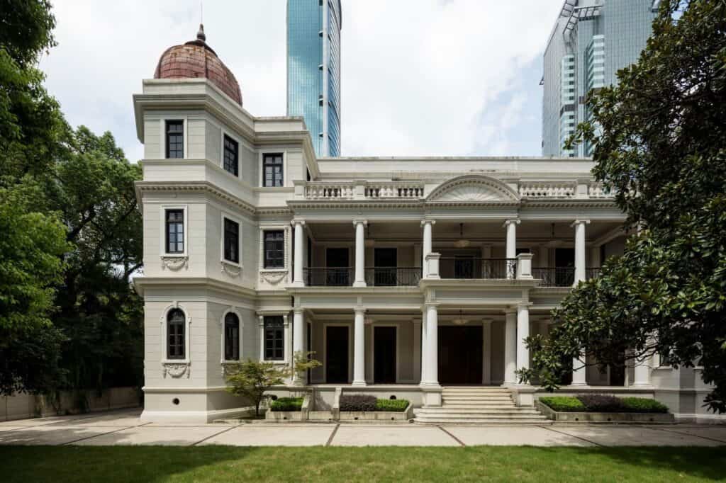 Daytime view of the garden facade of Prada Rong Zhai in Shanghai, a three-storey white villa with a red-domed corner tower, stacked columned loggias and a balustraded roof terrace above a lawn, with high-rise towers behind