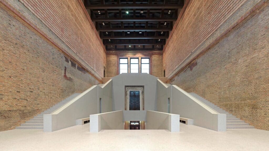 Symmetrical view of the white concrete staircase rising through the bare brick staircase hall of the Neues Museum to dark panelled doors in a stone surround, beneath three tall windows and a dark beamed ceiling