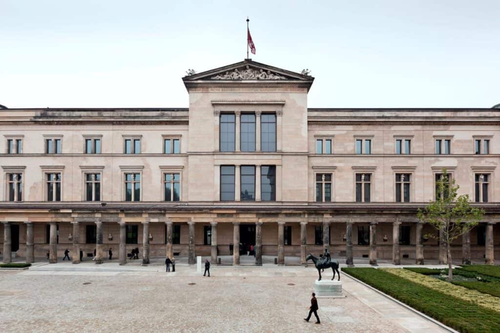East front of the Neues Museum with its pedimented central bay and long rows of windows above the restored stone colonnade, a bronze horse and rider standing on the paved forecourt