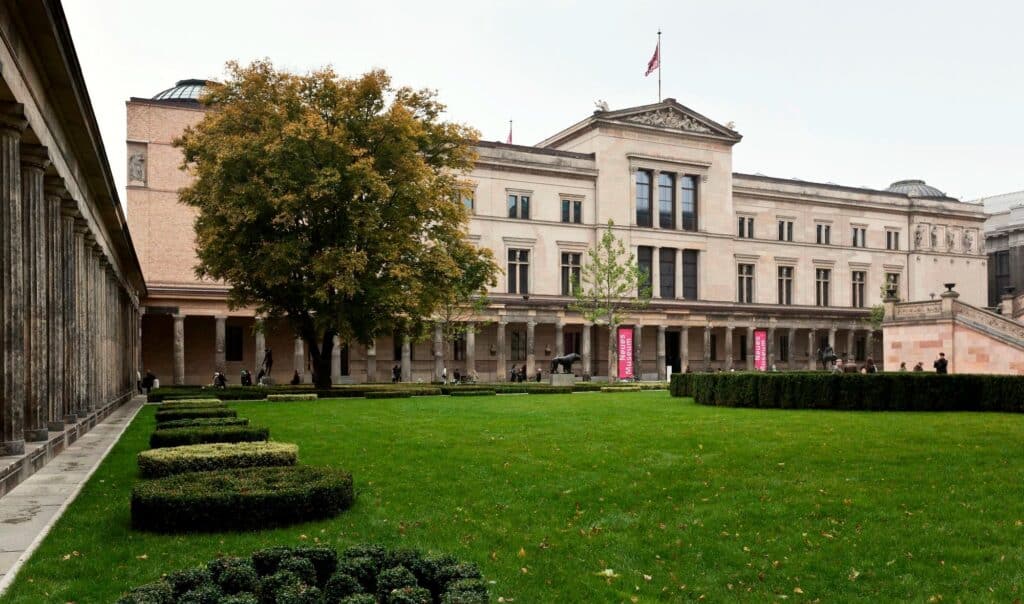Neues Museum seen across the lawn of the Colonnade Courtyard on Museum Island in autumn, framed by the stone colonnade and a broad tree, pink Neues Museum banners hanging between the columns