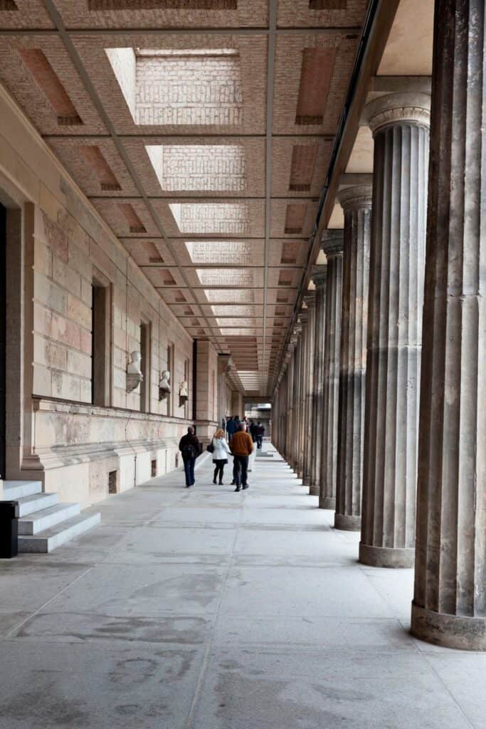 Long colonnade walk of the Neues Museum with fluted stone columns, a coffered ceiling pierced by skylights and busts set into the wall, visitors strolling into the distance