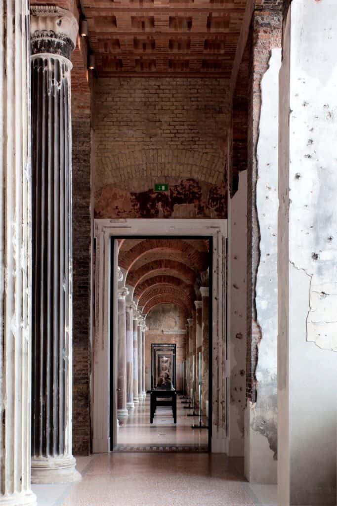 Enfilade through a doorway in the Neues Museum into a hall of brick vaults and columns lined with display cases, framed by a weathered fluted column and war-scarred plaster