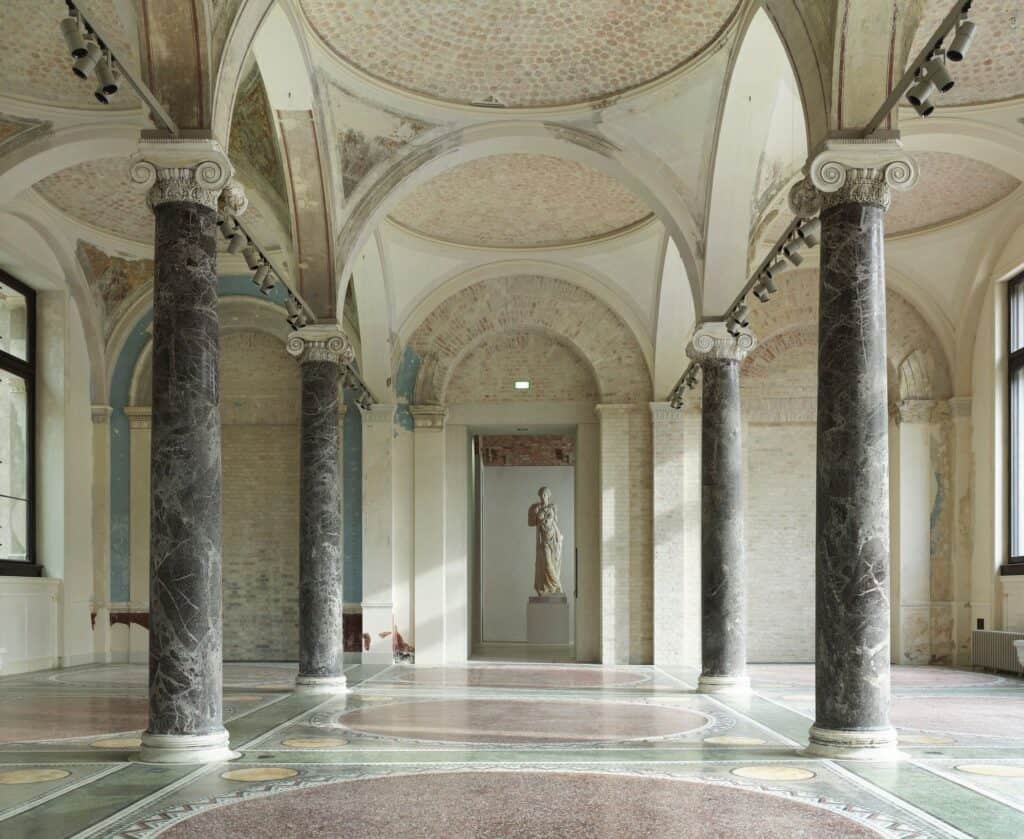 Restored hall in the Neues Museum with grey marble columns and Ionic capitals under shallow domed vaults, faded wall paintings and a patterned floor, a classical statue framed in the far doorway