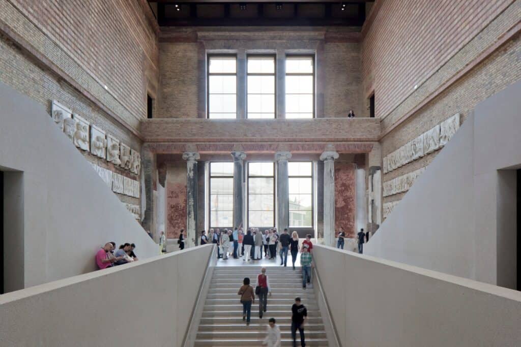 Visitors climbing the new white concrete staircase towards a screen of columns and tall windows in the brick staircase hall of the Neues Museum, relief panels lining the walls