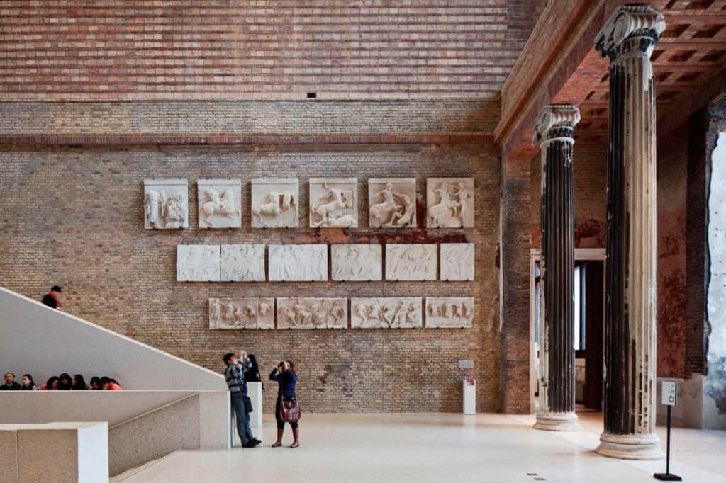 Rows of white relief panels on the bare brick wall of the Neues Museum staircase hall beside weathered fluted columns, visitors walking past the foot of the stair