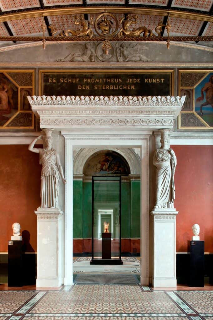 Ornate doorway flanked by caryatids beneath a gilded Prometheus inscription on a red wall in the Neues Museum, looking through to a bust in a tall glass vitrine