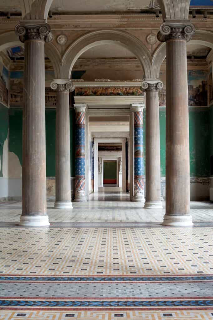 Enfilade of restored rooms in the Neues Museum framed by Ionic columns and painted blue and red columns, with green walls and a patterned mosaic floor