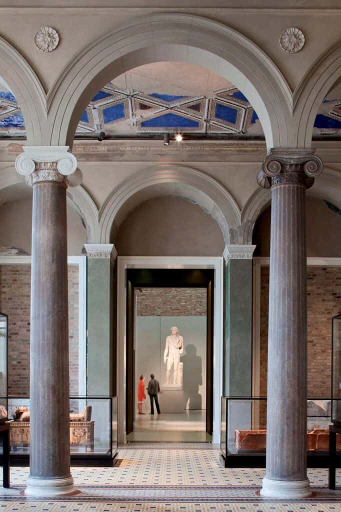 Pair of grey Ionic columns under arches and a blue coffered ceiling in the Neues Museum, framing a doorway to a white gallery with a classical statue and two visitors