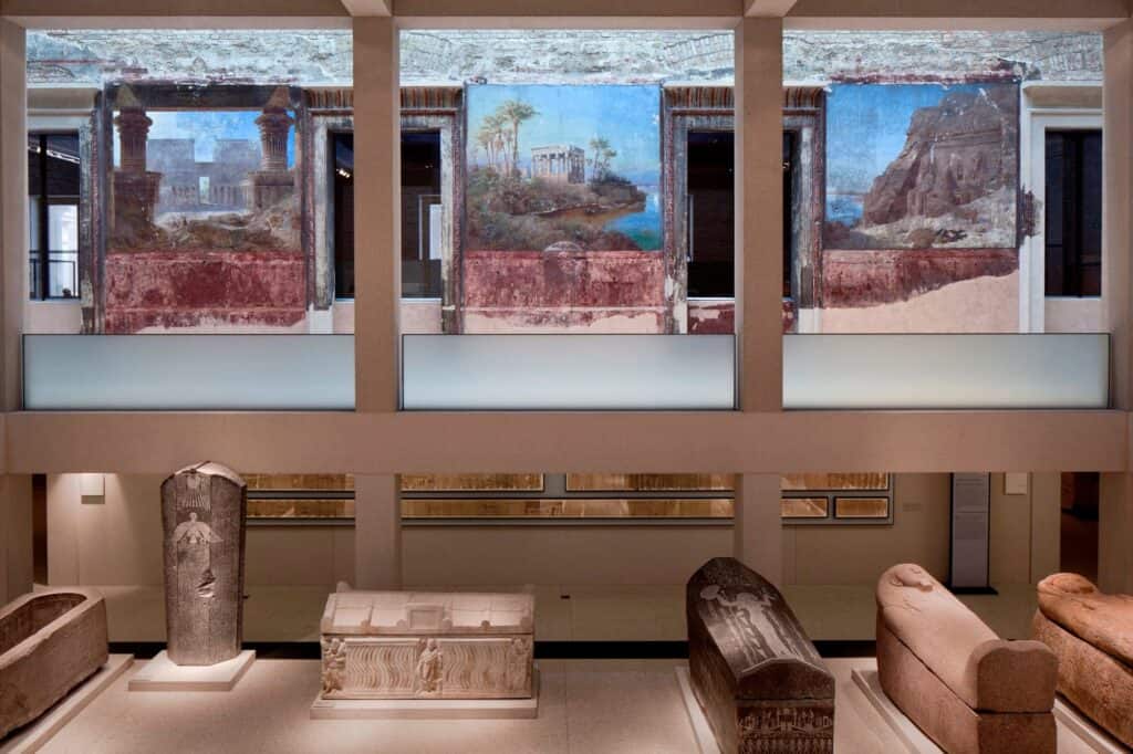 Egyptian Courtyard of the Neues Museum with surviving landscape wall paintings of temples above a concrete gallery frame, sarcophagi displayed on the floor below