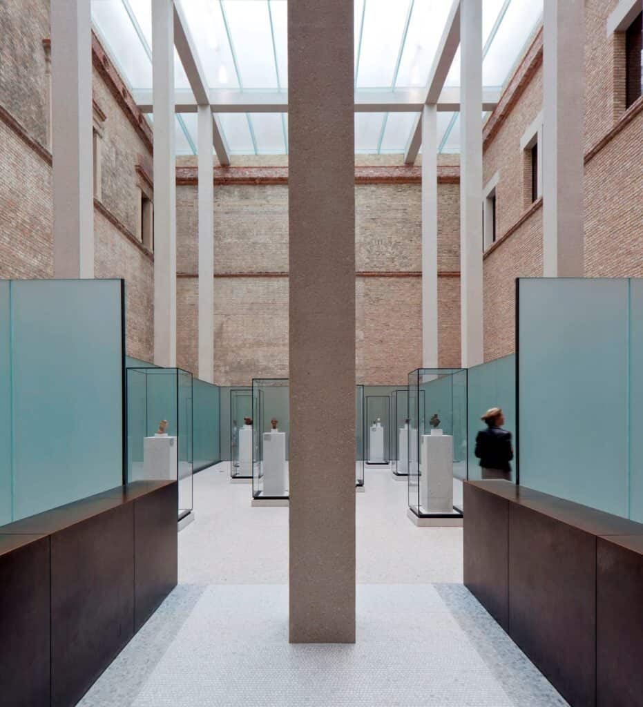 Glass vitrines of sculpted heads under the glazed roof of the Egyptian Courtyard at the Neues Museum, a square concrete column at the centre and brick walls beyond