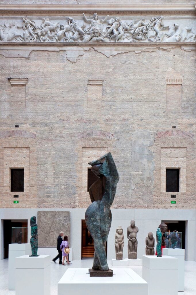 Bronze sculpture on a white plinth before the brick wall and sculpted frieze of the Greek Courtyard at the Neues Museum, stone figures and visitors behind
