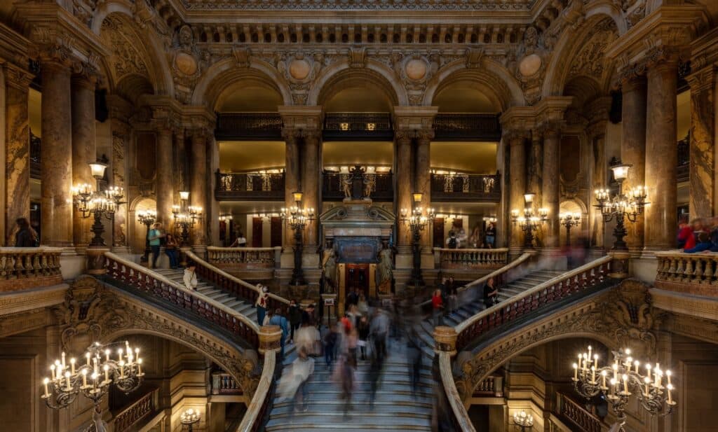 Grand staircase of the Palais Garnier in Paris seen from the upper landing, twin flights of white marble with red and green marble balustrades sweeping down between bronze candelabra as visitors blur on the steps