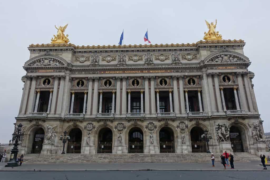 Main façade of the Palais Garnier on Place de l'Opéra in early morning, its arcaded loggia, paired columns and gilded corner sculptures topped by flags, with only a few people on the wide steps under a grey sky