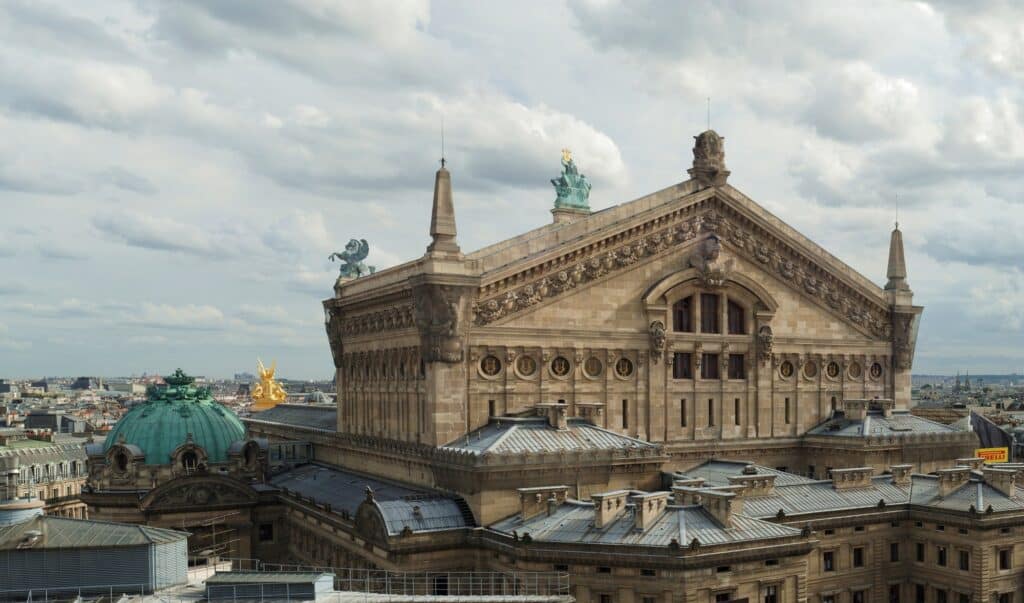 Rooftop view of the Palais Garnier, the tall stone gable of the stage house crowned by a bronze Apollo, with the green copper dome over the auditorium and the Paris rooftops under a cloudy sky