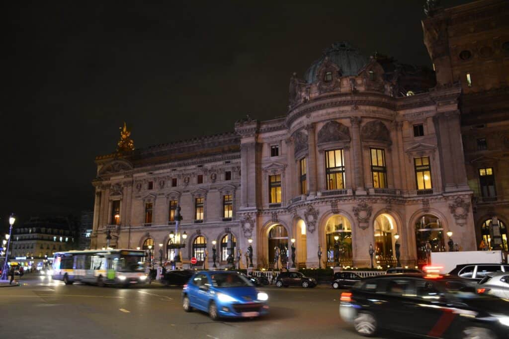 Side pavilion of the Palais Garnier lit at night, warm light on its arched windows and carved stone below a dark sky, as buses and cars streak past on the street