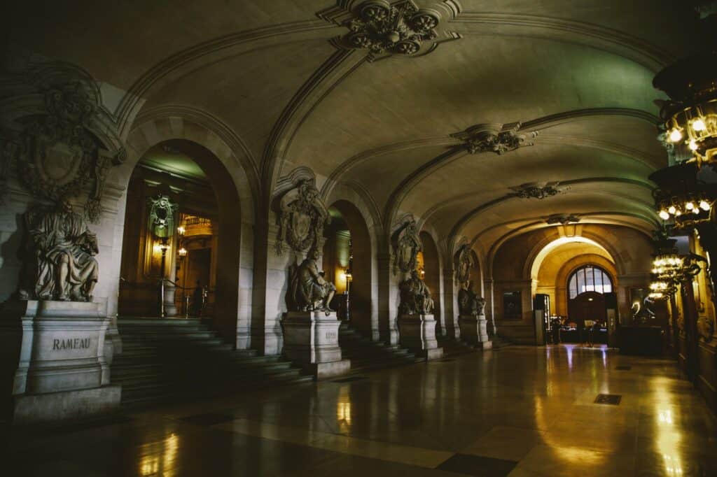 Vaulted lower vestibule of the Palais Garnier, stone statues on pedestals lining the arches and lanterns reflected in the polished floor leading towards the glowing staircase