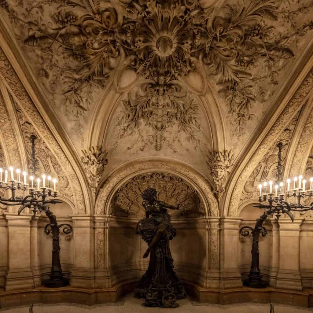 Bronze statue of the Pythia standing in a niche beneath an elaborately sculpted stone vault at the foot of the Palais Garnier staircase, flanked by tall candelabra