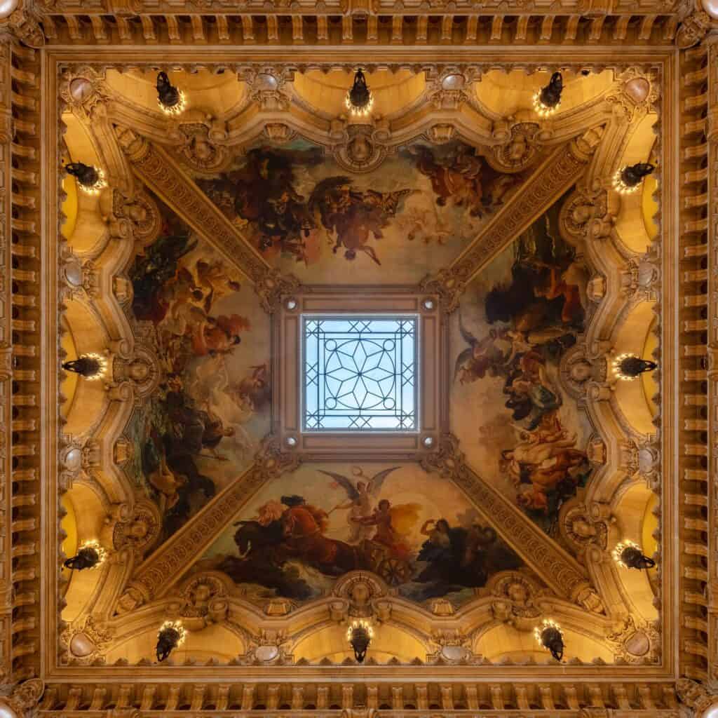Looking straight up at the ceiling of the grand staircase in the Palais Garnier, allegorical paintings framed in gilded mouldings around a central stained-glass skylight