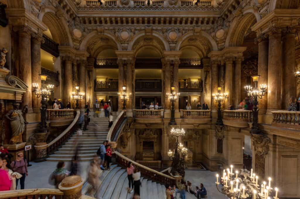 The grand staircase of the Palais Garnier seen from below, visitors climbing the marble steps beneath tiers of arcaded galleries, columns and clusters of lamps
