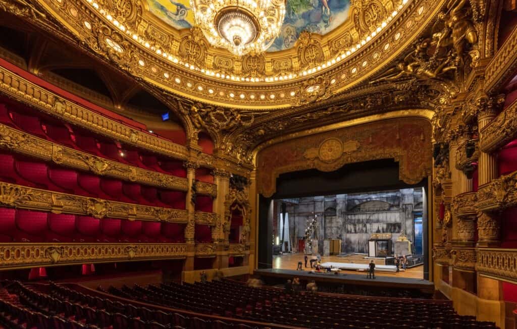 Horseshoe auditorium of the Palais Garnier seen from a side box, tiers of red velvet and gilded balconies curving towards a lit stage beneath the great chandelier