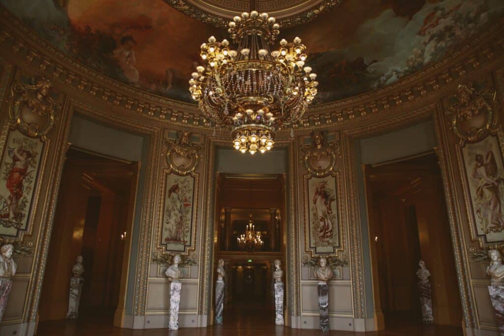Rotunda of the Salon du Glacier at the Palais Garnier, a large gilt and crystal chandelier hanging beneath a painted ceiling above tapestry panels, marble busts and tall doorways