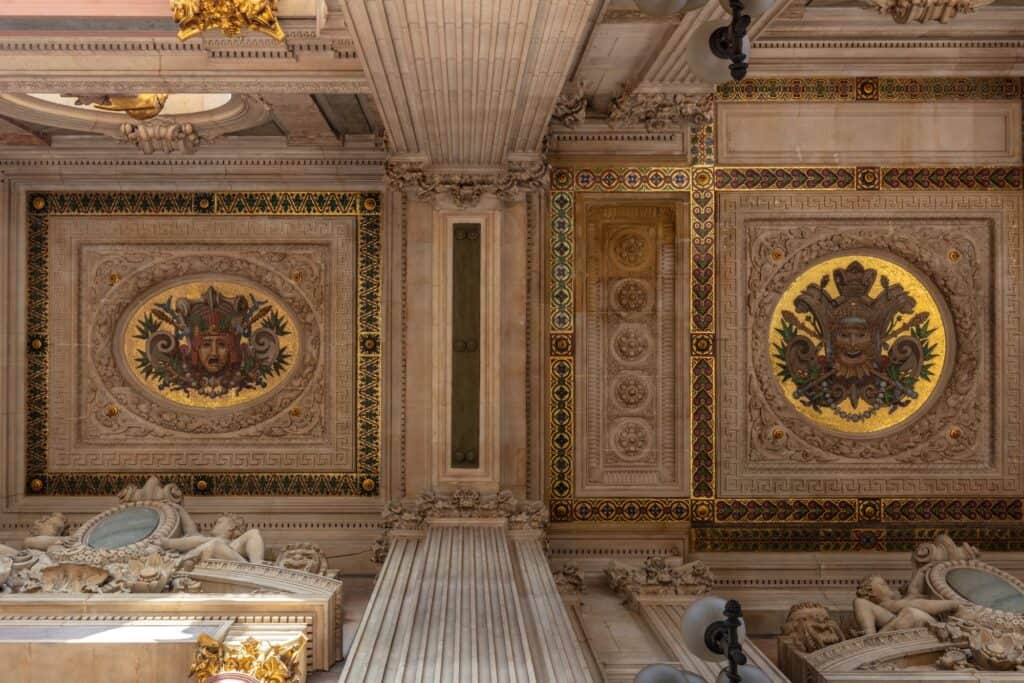 Coffered stone ceiling in an interior of the Palais Garnier, painted coats of arms in gilded roundels between marble cornices, with sculpted figures resting on the walls below