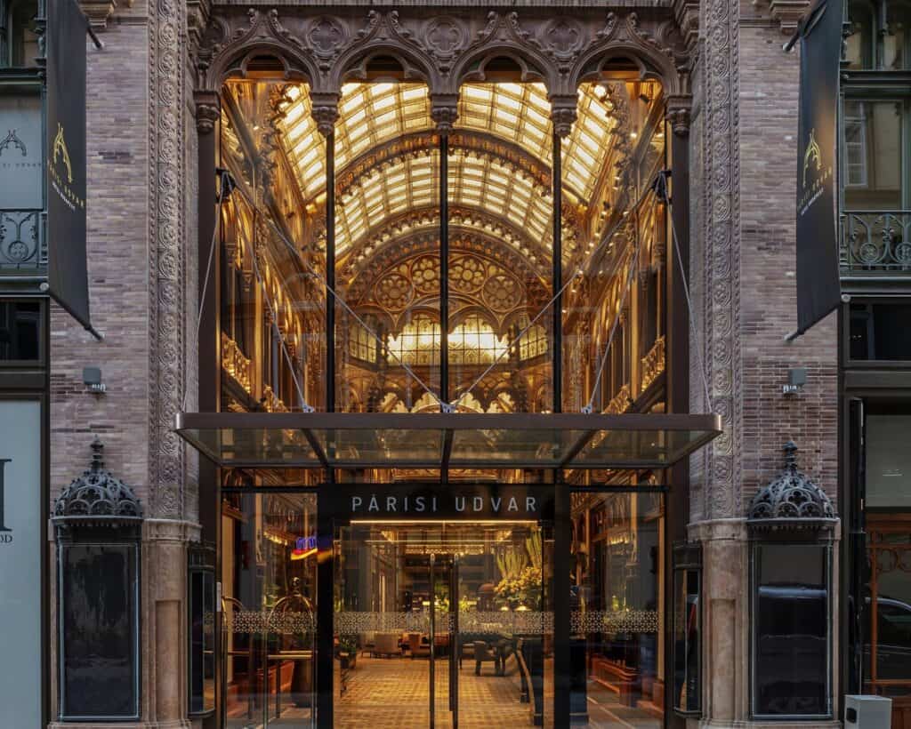 Street entrance of Paris Court on Ferenciek tere in Budapest, a tall glazed archway between carved brick piers revealing the golden glass vaults of the restored arcade, with a glass canopy over the Párisi Udvar doors