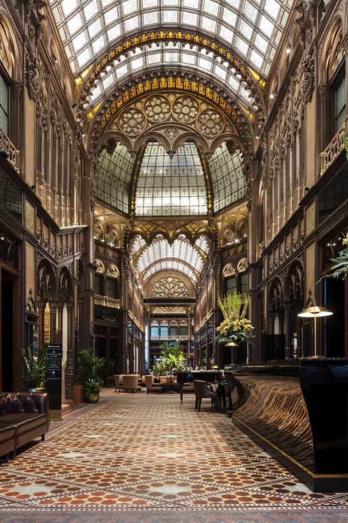 Long view down the restored Párisi Udvar passage, a patterned mosaic floor leading beneath tiers of carved dark timber and glowing glass barrel vaults, with potted palms and lamps along the way