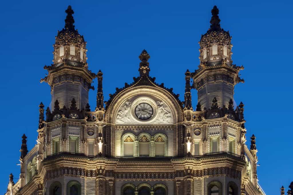 Twin turreted towers and the carved, floodlit crest of the Paris Court façade at dusk, ornate pinnacles, a round medallion and a row of arched windows set against a deep blue sky