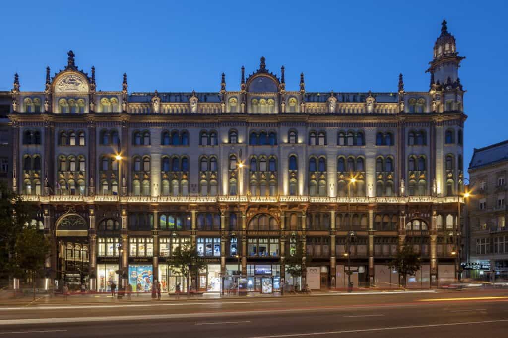 Full street front of the Párisi Udvar on Ferenciek tere at dusk, its long eclectic façade lit floor by floor above the shopfronts, with corner turrets and light trails on the road