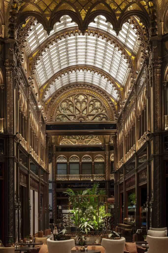 Hotel lounge in the restored Paris Court passage, armchairs and a potted tree at the foot of soaring carved arches, with a glass barrel vault and an ornate rose-patterned lunette beyond
