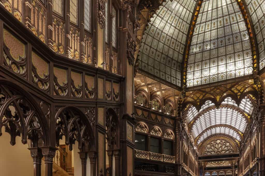 Richly carved timber arcading and pierced tracery around the upper galleries of the Párisi Udvar, rising to luminous glass vaults and a succession of ornamented arches