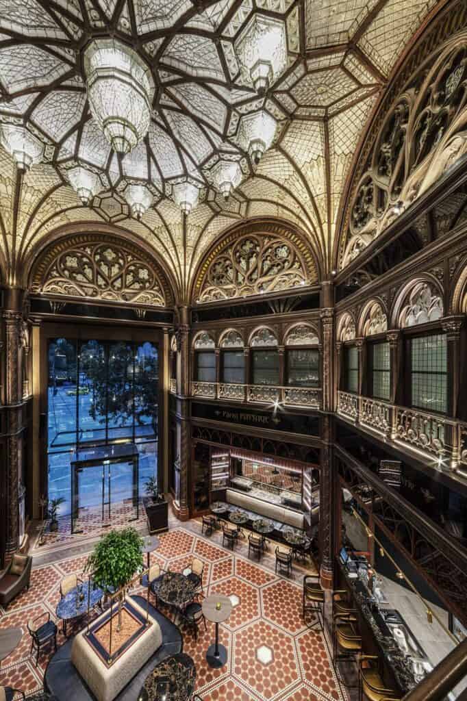 Wide view from an upper level of the Párisi Udvar, crystal chandeliers hanging from a dome of glass and gilded ribs above carved galleries, a glazed street entrance and a lobby lounge on a patterned floor